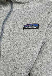 Grijze gebreide Patagonia trui met een kwart-ritssluiting aan de voorkant en een klein rechthoekig Patagonia logo op de borst.