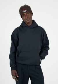 Granatowa bluza z kapturem z przednią kieszenią i prążkowanymi mankietami. Nosi się ją z ciemnymi spodniami oraz brązową czapką typu beanie z prążków z haftowanymi akcentami.