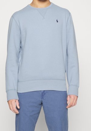 Person iført en ensfarvet lyseblå langærmet sweatshirt med et lille mørkt logo på venstre bryst og blå bukser.