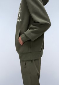 Sudadera verde oliva con bolsillo canguro, puños elásticos y dobladillo. Presenta un estampado gráfico en el pecho. Combinada con pantalones a juego.