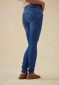 Uske skinny traperice u plavoj boji, s visokim strukom, suženim nogavicama, dvostrukim stražnjim džepovima i šivenim detaljima. Uparene s bež tkanim balerinama.