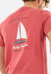 Camiseta de algodón roja con un gráfico de un velero. El texto incluye "Helly-Hansen" y "Vivo desde 1877", con un ajuste suave y casual.