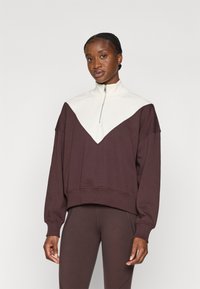 Brun og kremfarget hettejakke med glidelås og V-formet design, ribbestrikkede mansjetter og myk tekstur. Kombinert med matchende brune leggings.