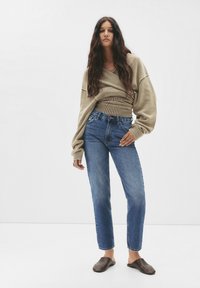 Beige oversized trui met geribbelde onderrand, blauwe rechte jeans en bruine instapschoenen. De textuur lijkt zacht; het ontwerp is casual.