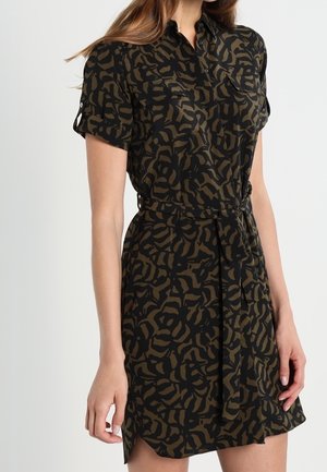 Vrouw draagt een kortmouwige, met riem gesmokte overhemdjurk met een zwart-olijfgroen abstract dierenprintpatroon.