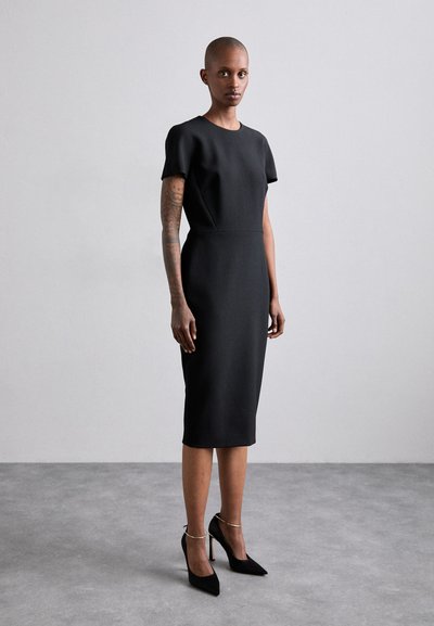 Vestito nero al ginocchio con maniche corte, tessuto strutturato, scollatura rotonda e vita aderente. Abbinato a tacchi neri e dettagli in oro.