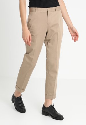 Beige slim-fit broek met omslag, gedragen met zwarte leren veterdressen en een zwart geribd mouwloos topje.
