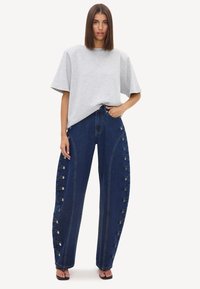 Femme portant un t-shirt oversize gris clair et un jean large bleu foncé avec des détails de boutons le long des jambes extérieures, debout avec une main dans la poche.