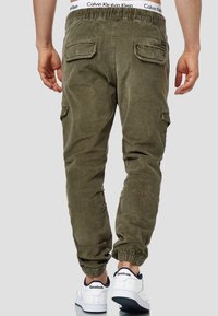 Zielone khaki spodnie cargo z elastycznym pasem i mankietami, z wieloma kieszeniami i teksturowanym materiałem, noszone z białymi sneakersami.