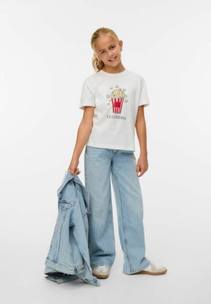 Ragazza giovane che sorride, indossa una maglietta bianca con grafica di popcorn e testo "Le Cinéma", jeans larghi, tiene una giacca di jeans in una mano.