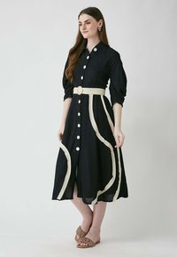 Ein marineblaues Knopfkleid mit tailliertem Gürtel und weißen, geschwungenen Akzenten. Hergestellt aus einem strukturierten Stoff, hat es kurze, puffartige Ärmel.