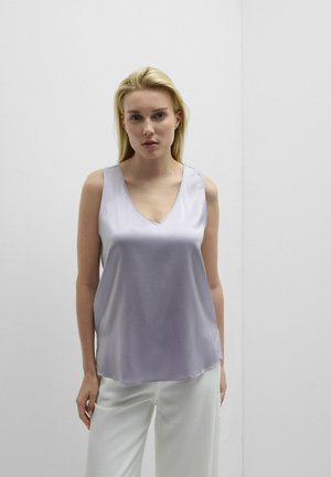 Blonde vrouw met een mouwloze lavendelkleurige satijnen top en witte broek, staande tegen een effen lichtgrijze muur.