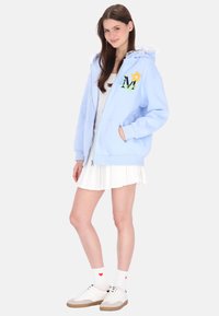 Hellblauer Kapuzenpullover mit Reißverschluss, gerüschter Kapuze, floraler Stickerei und zwei Fronttaschen. Gestylt mit einem weißen Rock und weißen Turnschuhen.