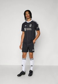 Equipamento de futebol preto com camisola de manga curta com detalhes brancos, calções combinando e meias brancas até ao joelho com riscas pretas.