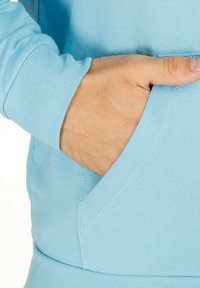 Felpa blu chiaro con cappuccio, dotata di tasca, texture morbida e polsini a coste. Il design presenta cuciture in vista e una vestibilità comoda.