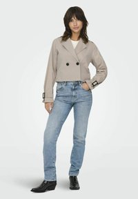 Veste beige cintrée avec un design à double boutonnage, boutons noirs et manches larges, associée à un jean bleu clair et des bottines à talons noires.