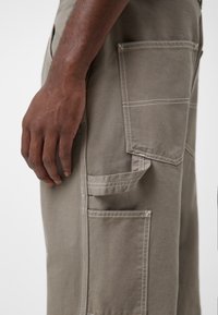 Světle šedé cargo kalhoty se rovný střihem, které mají více náprsních kapes a kontrastní švy. Materiál vypadá odolně a texturovaně.