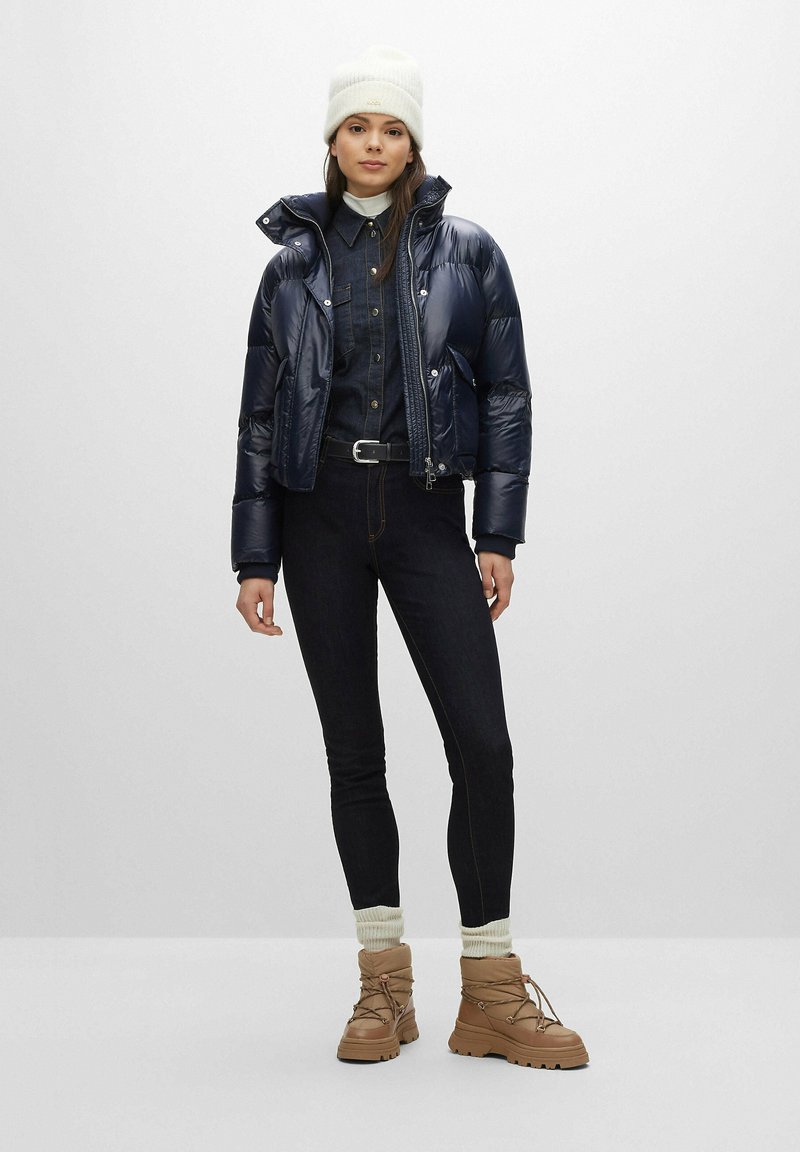 Chaqueta de plumas azul marino, con alto cuello, combinada con una camisa de mezclilla, jeans negros y botas beige con cordones. Un gorro de punto beige completa el look.