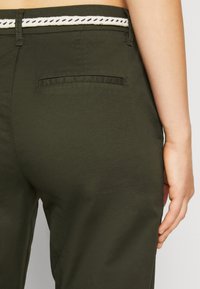 Pantalons vert kaki avec une texture lisse, dotés d'une ceinture tissée blanche et bleu marine en accent et d'un détail de poche arrière.