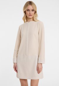 Beiges Shiftkleid mit hohem Kragen, langen Ärmeln, strukturierten Rippakzenten und einem glatten oberen Abschnitt mit einer mittigen Naht.