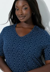 Mujer sonriente con cabello largo y oscuro, que lleva pendientes de aro dorados y una camisa azul marino con patrones circulares blancos sobre un fondo claro.