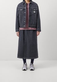 Denimjas met verticale witte strepen, knopen aan de voorkant, en twee borstzakken; gecombineerd met een donker gestreepte lange rok en sneakers.