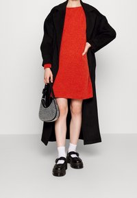 Abrigo largo negro, vestido de punto rojo, bolso plateado con embellishments, calcetines blancos y zapatos de plataforma negros con detalles de hebilla.