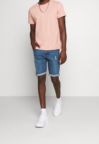 Un t-shirt rose clair à manches courtes associé à un short en denim bleu clair avec des ourlets roulés et des détails déchirés, complété par des baskets blanches.