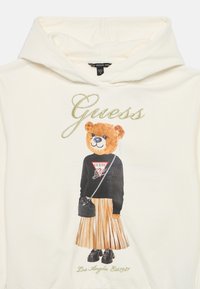 Felpa crema con un grafico di un orso in un maglione nero, una gonna plissettata dorata e scarpe nere, con il testo "Guess" e decorazioni.