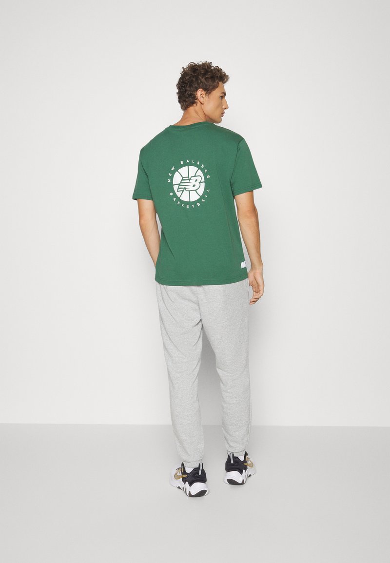 Grünes Baumwoll-T-Shirt mit weißem Logo-Druck auf der Rückseite, kombiniert mit hellgrauen Jogginghosen und schwarz-goldenen Sneakern.