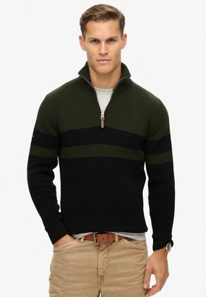 Strikket pullover i mørkegrøn og sort med lynlåskrave. Har ribbet tekstur og vandrette striber. Bæres over en grå skjorte.
