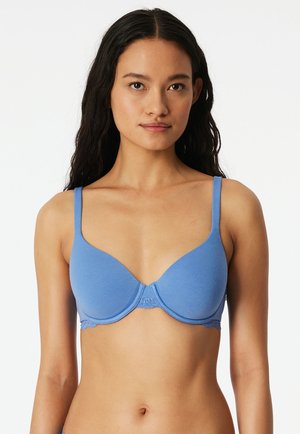 Junge Frau mit langen dunklen Haaren, die einen hellblauen Bügel-BH mit dünnen Trägern trägt, steht vor einem weißen Hintergrund.