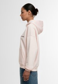 Rosa bomullshettegenser med frontlomme, justerbar snorhette og brodert tekst. Løs passform, glatt tekstur og ribbede mansjetter.