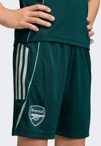 Pantalones cortos deportivos verde oscuro con detalles en azul claro, que cuentan con un logo blanco de Arsenal y tres rayas verticales blancas en un lado.