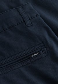 Tamsiai mėlynos audinio kelnės kišenė su maža juoda GABBA etiketė, siūta netoli skeltos kišenės angos.