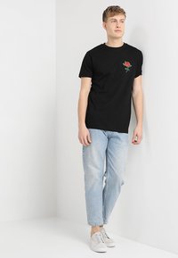 Camiseta de algodón negra con un pequeño bordado de rosa roja en el lado izquierdo del pecho, combinada con jeans de mezclilla azul claros y zapatillas blancas.