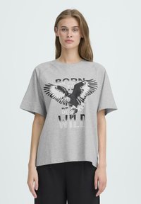 Camiseta gris de gran tamaño hecha de algodón con un estampado gráfico de un águila y el texto "NACIDO PARA SER SALVAJE". Presenta mangas cortas y un corte relajado.