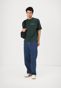 Zaļš kokvilnas T-krekls ar baltu tekstu, apvienots ar brīvām zilām džinsa biksēm un baltām kurpēm ar sarkanām svītrām. Melna plecu soma.