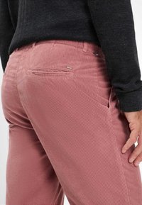 Pantaloni in velluto a coste rosa con una texture rigata, tasche laterali e un piccolo logo "Brax" sulla tasca posteriore, abbinati a un maglione scuro.