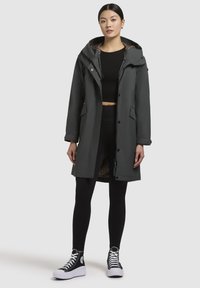 Parka gris foncé avec capuche, présentant une doublure léopard, des boutons sur le devant et deux poches latérales. Portée par-dessus des leggings noirs et des chaussures.