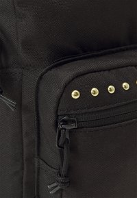 Sac en tissu noir offrant une poche zippée, des clous décoratifs dorés et une surface texturée. Comprend du matériel noir et un cordon réglable.