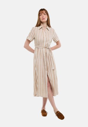 Femme debout, mains derrière le dos, portant une robe midi beige à rayures blanches verticales avec col et chaussures plates marron à enfiler.