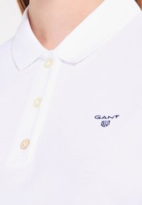 Hvid polo-skjorte lavet af struktureret stof, med en klassisk krave, tre knapper og et lille blåt "GANT" logo på venstre bryst.
