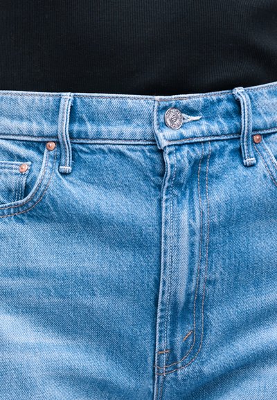 Φαρδιά τζιν ανοιχτού μπλε με κουμπί και θηλιές για ζώνη, φορεμένα με μαύρο ριμπ μπλουζάκι που έχει μπει μέσα.