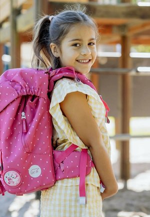 Junges Mädchen, das draußen lächelt, trägt ein gelbes karierte Kleid und einen großen rosa Rucksack mit weißen Punkten.