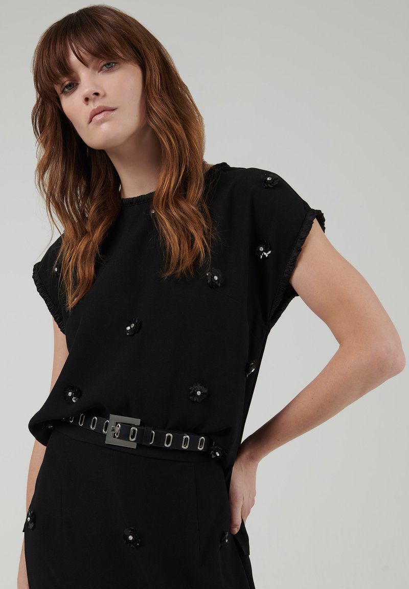 Top negro de manga corta, con embellishments florales texturizados y un escote con volantes. Combina con un cinturón negro y una silueta elegante.