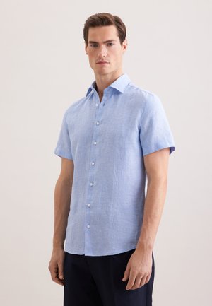 Mężczyzna w jasnoniebieskiej koszuli z krótkim rękawem na guziki i ciemnych spodniach, stojący na tle jednolitego jasnego tła, zwrócony przodem.