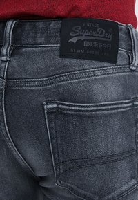Vista trasera de un bolsillo de jeans de mezclilla gris oscuro con la etiqueta de la marca SuperDry en negro y tela roja visible sobre la cinturilla.