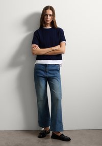Top de punto de manga corta azul marino con textura, superpuesto sobre una camisa blanca, combinado con unos vaqueros de pierna ancha de un azul claro y mocasines negros.