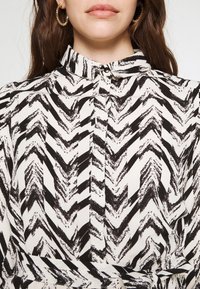 Black and white patterned blouse featuring a chevron design, collared neckline, and button closure at the front. Textured fabric.